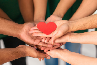 A group of hands from different individuals holding a red heart cutout, representing community support and care.