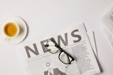 A close-up of a newspaper with the word "NEWS" in bold, a pair of glasses, and a cup of coffee on a white surface.