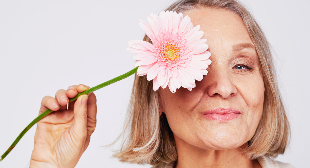 A confident older woman with short blonde hair holds a pink gerbera daisy over one eye, smiling gently. She has a serene expression, and the background is a clean, neutral white.