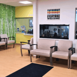 Bright and welcoming pediatric waiting room featuring a birch tree mural, soft beige chairs with black armrests, a wall-mounted aquarium, and a “Everyone Welcome” sign, creating a calming and inclusive environment for children and families.