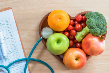 A wooden bowl with fresh fruits and vegetables arranged in the shape of a heart, with a stethoscope placed beside it.