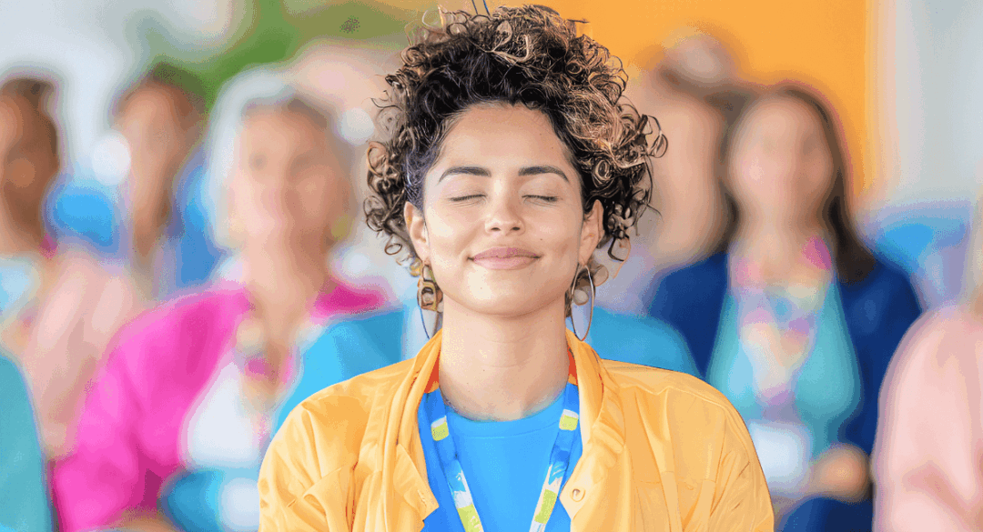 A young woman with curly hair and a relaxed expression practices mindfulness meditation with her eyes closed in a group setting. The background shows a diverse group of people engaged in the same activity, emphasizing stress management and emotional well-being.