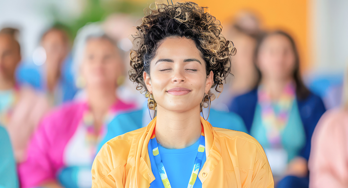 A young woman with curly hair and a relaxed expression practices mindfulness meditation with her eyes closed in a group setting. The background shows a diverse group of people engaged in the same activity, emphasizing stress management and emotional well-being.