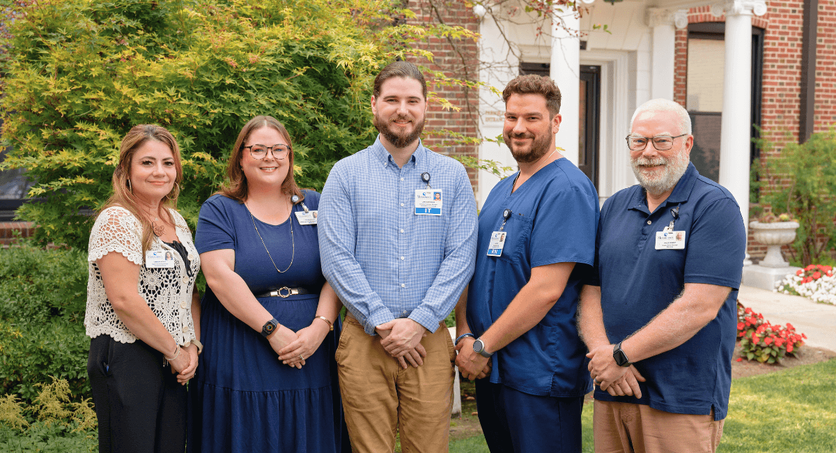 Five Monadnock Community Hospital employees stand together outside the hospital, smiling and wearing professional attire, representing gratitude for staff, volunteers, donors, and the community.
