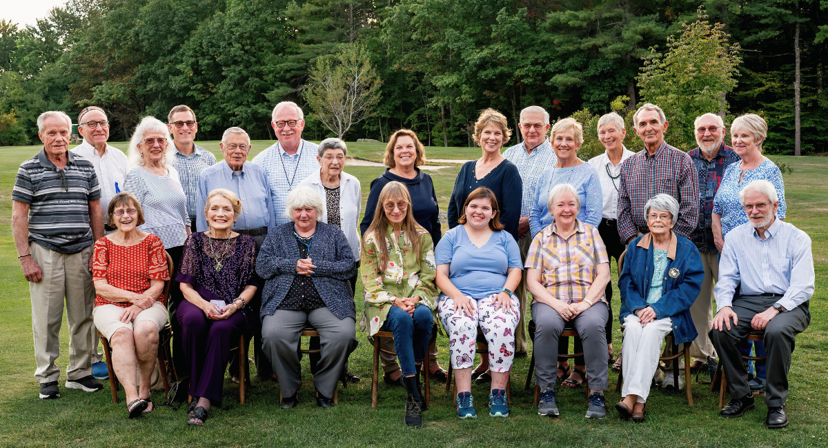 Group photo of 26 Monadnock Community Hospital volunteers outdoors, smiling warmly. The image includes a mix of seated and standing individuals with a forested background.