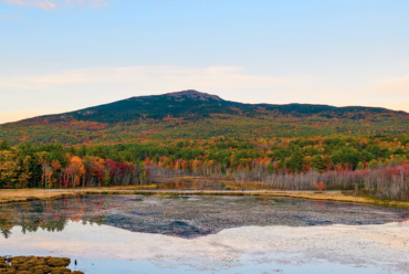 A scenic view of Mount Monadnock reflected in a calm pond, surrounded by colorful autumn foliage under a soft blue sky.