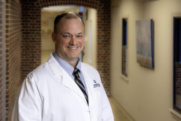 Dr. Michael Gilbert, wearing a white coat and striped tie, smiles while standing in a warmly lit hallway with brick accents and framed artwork on the walls.