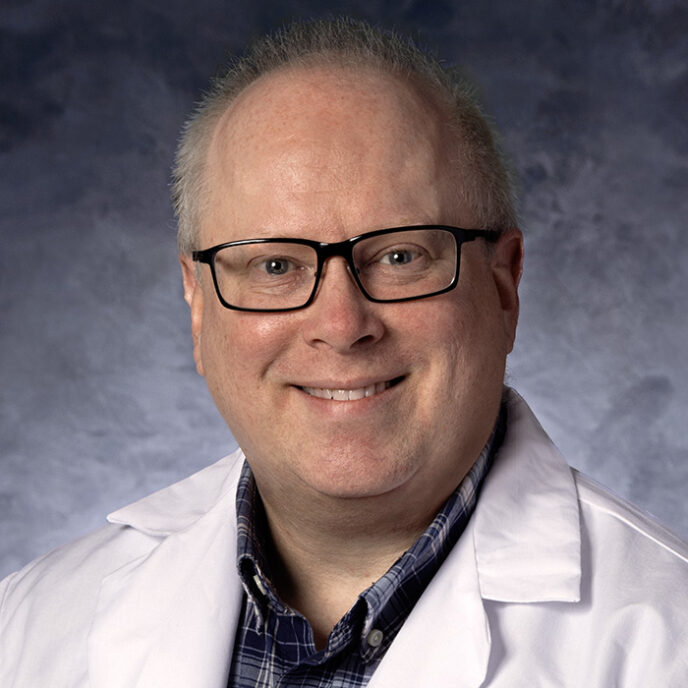 A smiling healthcare professional wearing glasses, a plaid shirt, and a white lab coat, posed in front of a softly textured blue-gray background.