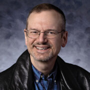 A smiling man wearing wire-rimmed glasses, a black leather jacket, and a blue plaid shirt, posed in front of a textured blue-gray background.