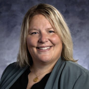 A smiling woman with blonde hair wearing a dark green blazer and a gold pendant necklace, posed against a softly lit blue-gray background.