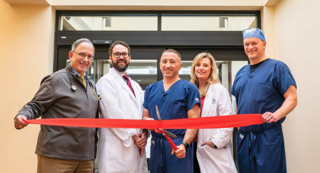 Five Monadnock Community Hospital providers smile while holding a red ribbon across a sunlit corridor under a skylight. From left to right: Dr. Jonathan Krant, Dr. Robert Spencer, Dr. Vache Hambardzumyan, APRN Brooke O’Connor, and Dr. Michael Gilbert.