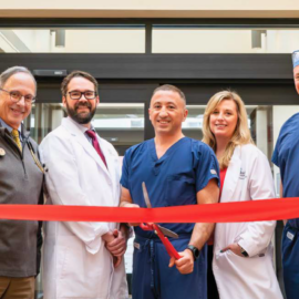Five Monadnock Community Hospital providers smile while holding a red ribbon across a sunlit corridor under a skylight. From left to right: Dr. Jonathan Krant, Dr. Robert Spencer, Dr. Vache Hambardzumyan, APRN Brooke O’Connor, and Dr. Michael Gilbert.