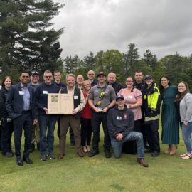A large group of EMS professionals, MCH staff, and supporters gather on a golf course lawn, smiling around Captain Tim Quinn who holds the Lauren Morton Award.