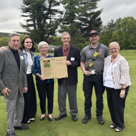 Members of Lauren Morton's family stand with Captain Tim Quinn on the grass, proudly holding a tribute plaque next to him and smiling for the camera.