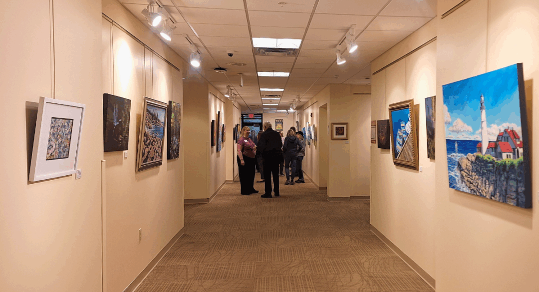 A hallway filled with colorful paintings on both walls during an art exhibition at Monadnock Community Hospital. Several visitors are gathered and chatting in the distance beneath gallery lights.