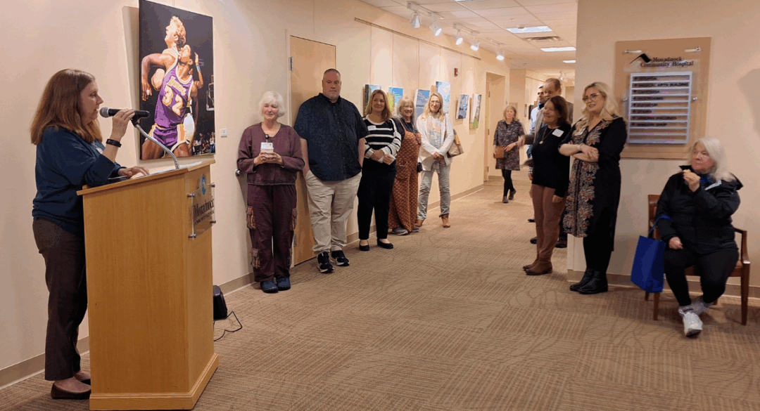 Katharyn speaks at a podium with a microphone in front of a group of people at an art gallery opening. The group stands along the hallway wall, listening attentively. Behind her is a painting of two basketball players. A wall sign for Monadnock Community Hospital is visible.