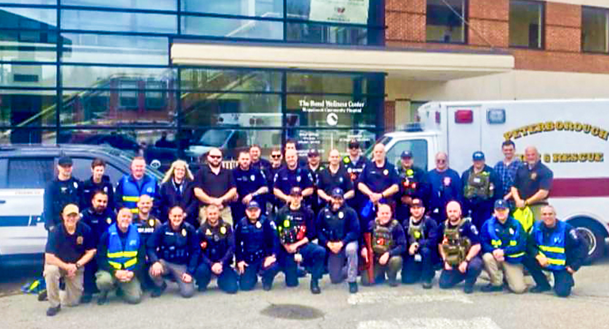 A large group of first-responders—including police officers, EMS, and fire personnel—pose together in uniform outside the Bond Wellness Center at Monadnock Community Hospital. Emergency vehicles are parked behind them, including a Peterborough Fire & Rescue ambulance.
