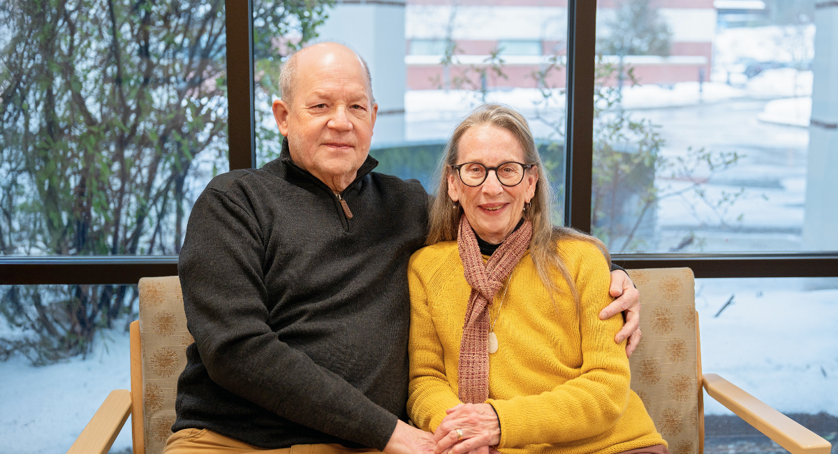 Craig and Judy Unger-Clark sit together smiling in a hospital waiting area with snow outside the window. Craig wears a dark sweater; Judy is in a yellow sweater and scarf.