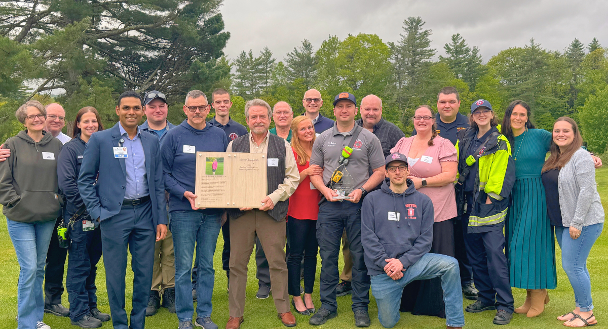 A group of more than a dozen EMS professionals, MCH staff, and family, posing on a lawn with Captain Tim Quinn holding an award, surrounded by smiling colleagues and loved ones.