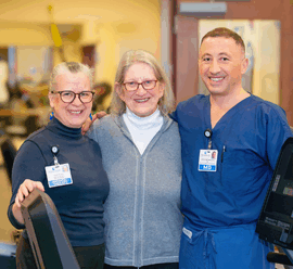 Rosamond Cady stands between two smiling healthcare providers—one woman in glasses and a black turtleneck with a name badge, and one man in blue scrubs with an MD badge. They are in a bright, welcoming rehabilitation or wellness area with fitness equipment visible in the background.