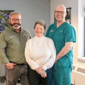 Fontaine Melus stands smiling between two members of the Mobile Integrated Healthcare team from Monadnock Community Hospital. On the left is Dr. Lucas Shippee in a green shirt, and on the right is paramedic Joshua Patrick in green scrubs.