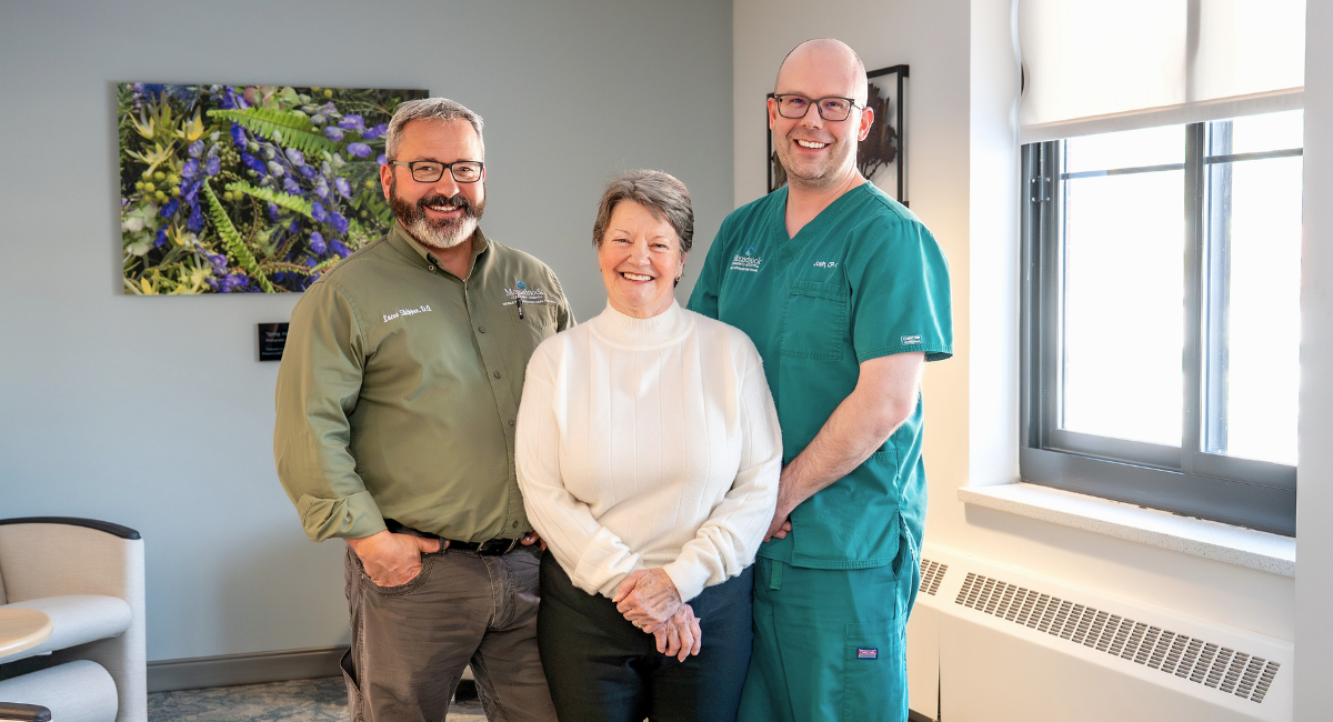 Fontaine Melus stands smiling between two members of the Mobile Integrated Healthcare team from Monadnock Community Hospital. On the left is Dr. Lucas Shippee in a green shirt, and on the right is paramedic Joshua Patrick in green scrubs.