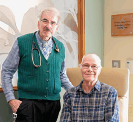 An older man in a green sweater vest with a stethoscope stands beside a seated patient, Don Nalette, who is wearing a plaid shirt and smiling. They are in a warmly lit exam room with a floral wall design and a dedication plaque in the background.