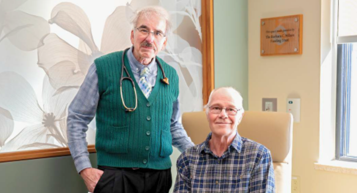 Dr. Eric Bonnem stands beside patient Don Nalette in a bright, welcoming infusion therapy room at Monadnock Community Hospital. Don is seated and smiling, wearing a plaid shirt, while Dr. Bonnem wears a green sweater vest with a stethoscope.