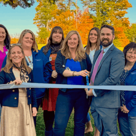 The Monadnock Cardiology Associates team stands outside in front of colorful fall trees, smiling while holding a silver ribbon for their ribbon-cutting ceremony.