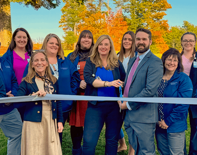 The Monadnock Cardiology Associates team stands outside in front of colorful fall trees, smiling while holding a silver ribbon for their ribbon-cutting ceremony.