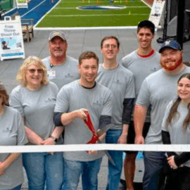 The Monadnock Community Hospital rehabilitation team stands together outdoors, smiling and holding a large ribbon in front of the new outdoor turf rehabilitation space. A staff member at the center holds oversized scissors. The turf area in the background features agility cones and a Free Throw Shoot Out sign, emphasizing a fun and functional therapy environment.