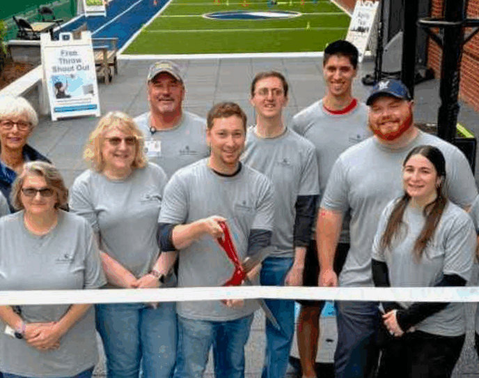 The Monadnock Community Hospital rehabilitation team stands together outdoors, smiling and holding a large ribbon in front of the new outdoor turf rehabilitation space. A staff member at the center holds oversized scissors. The turf area in the background features agility cones and a Free Throw Shoot Out sign, emphasizing a fun and functional therapy environment.