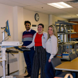 Three administrative staff members standing by a computer workstation in an office with shelves and supply boxes in the background.