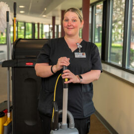 Environmental Services staff member in black scrubs stands with a vacuum and cleaning cart in a sunny hospital hallway.