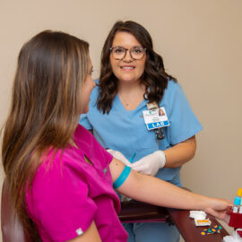 Lab technician in blue scrubs smiles while drawing blood from a patient in a pink top during a phlebotomy procedure.
