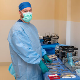 Surgical technician in full sterile gown and gloves stands beside a table of surgical instruments in a brightly lit procedure room.
