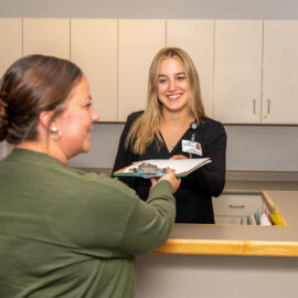 Office assistant hands a clipboard to a patient at a medical reception counter, both smiling during the exchange.