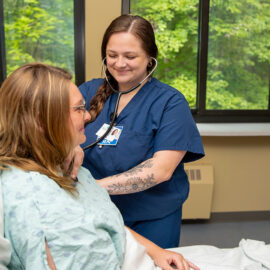 Registered nurse in navy scrubs listens to a patient’s heartbeat with a stethoscope in a hospital room with large windows.