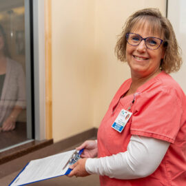 Emergency department registrar in coral scrubs holds a clipboard and smiles while assisting a patient at a registration window.