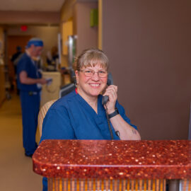 Health Unit Coordinator in navy scrubs answers a phone at a hospital workstation with clinical staff visible in the background.