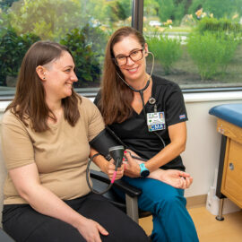 Certified medical assistant in black scrubs takes a patient’s blood pressure in a sunny outpatient exam room.