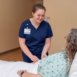 A Licensed Nursing Assistant (LNA) in navy scrubs gently assists a patient in a hospital bed, both smiling and engaged in conversation.