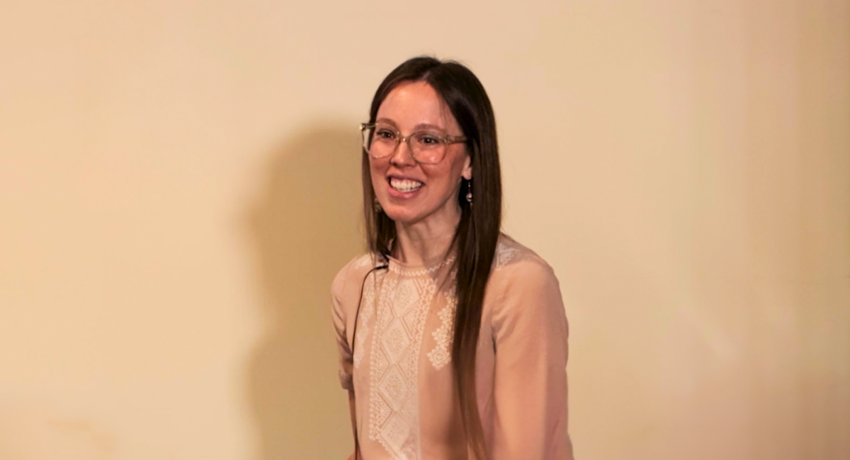 Dr. Elizabeth Johnsen smiles while speaking in front of a neutral beige background, wearing glasses and a light pink blouse with lace detailing.
