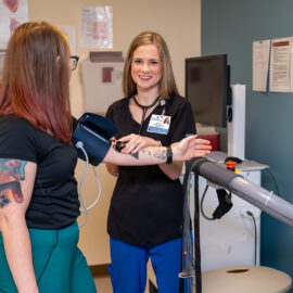 A cardiology technician in black scrubs checks a patient’s blood pressure while smiling toward the camera, inside a cardiology exam room.