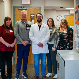 Five staff members from the cardiology team, including a provider in a white coat, stand together and smile in a clinical hallway.