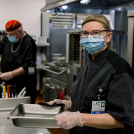 A masked food service worker in black attire holds a pan and smiles in a hospital kitchen while a coworker prepares food in the background.