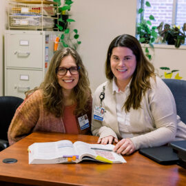 Two hospital dietitians sit side by side at a desk, smiling, with an open nutrition book in front of them in a shared office space.