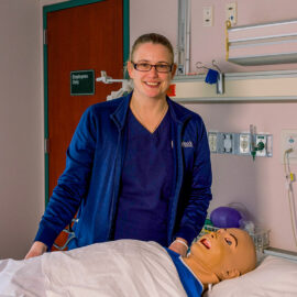 A nurse educator smiles while standing beside a medical training mannequin in a hospital simulation room.
