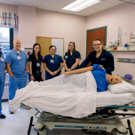 A team of nurses and educators pose around a hospital bed with a training mannequin in a clinical simulation space.