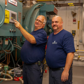 Two members of the facilities and engineering team stand next to boiler controls and piping in the hospital’s mechanical room, smiling.